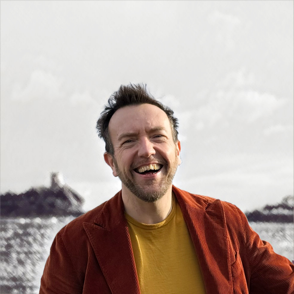 A smiling middle aged white male with short mousey hair and a close cropped beard stands facing camera wearing a mustard jumper under a rust coloured corduroy jacket.