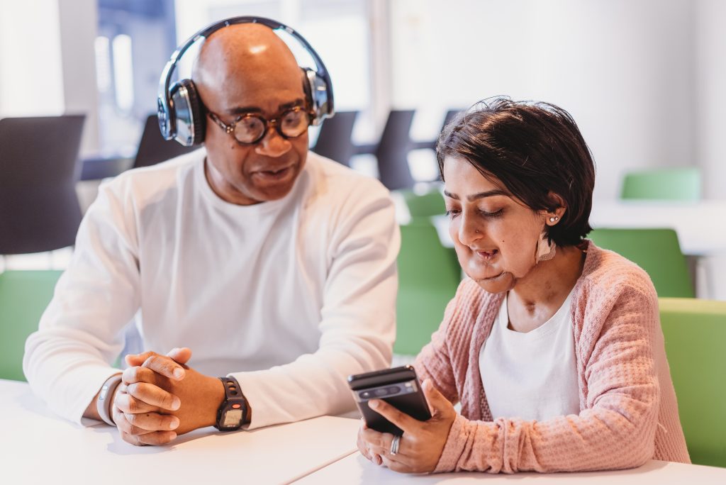 Person who is short in stature and with a facial difference looks at phone with friend or colleague who is wearing headphones