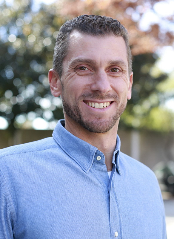 A middle aged white male wearing a blue button down collar shirt. He has brown eyes, short curly brown hair and a close cropped beard. he is looking towards camera smiling.