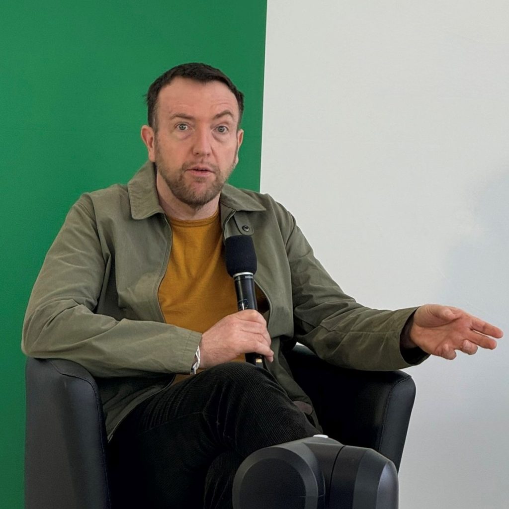 A seated man speaking into a handheld microphone at what appears to be a talk or interview event. He has light skin, short dark hair, and a short beard, and he is sitting in a black armchair with one leg crossed, gesturing with one hand while holding the microphone with the other. He is wearing a light olive-green jacket over a mustard-yellow shirt, dark pants, and dark shoes. Behind him is a large backdrop split into green (left) and white (right). In the foreground, a large black camera or recording device with cables is positioned in front of him, partly blocking the lower part of the scene.